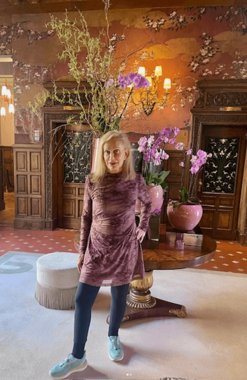 About Linda Rickels: A woman in a purple dress and blue sneakers stands in front of a round table with pink vases and orchids, in an ornately decorated room with wood paneling and floral wallpaper.