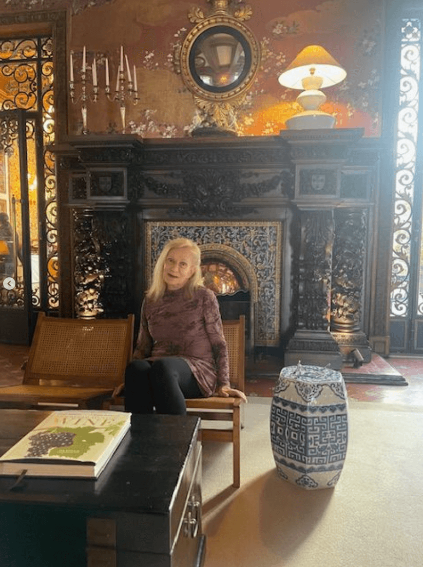 An older woman sits on a wooden bench in an ornate room with a decorative fireplace, intricate tiles, a lamp, and a patterned ceramic stool.